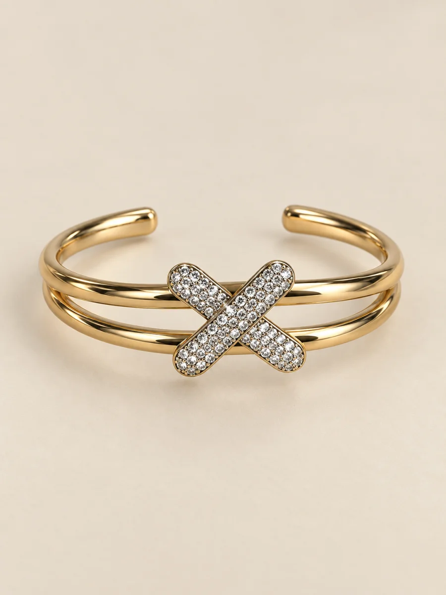 Bracelete dourado com X cravejado sobre fundo bege