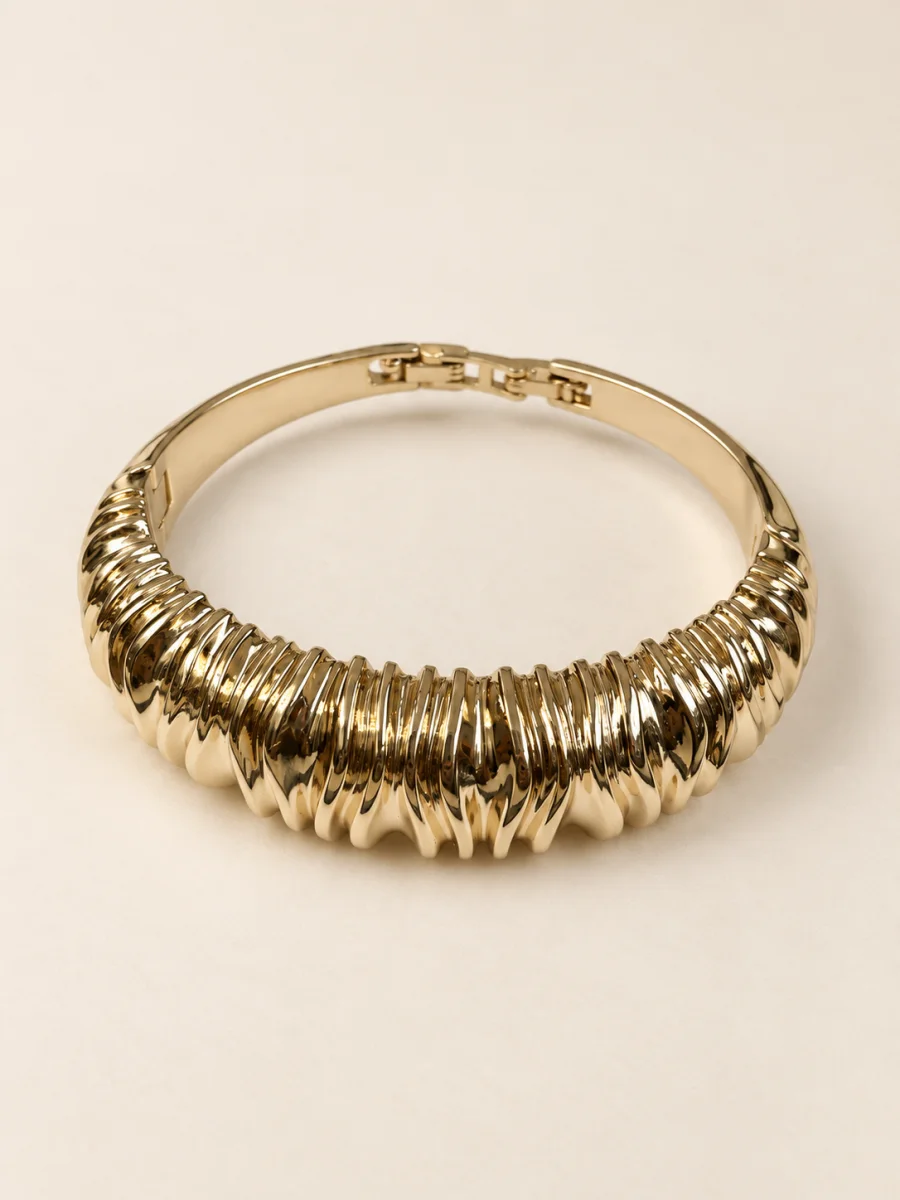 Pulseira dourada rígida com frente plissada sobre fundo bege claro.