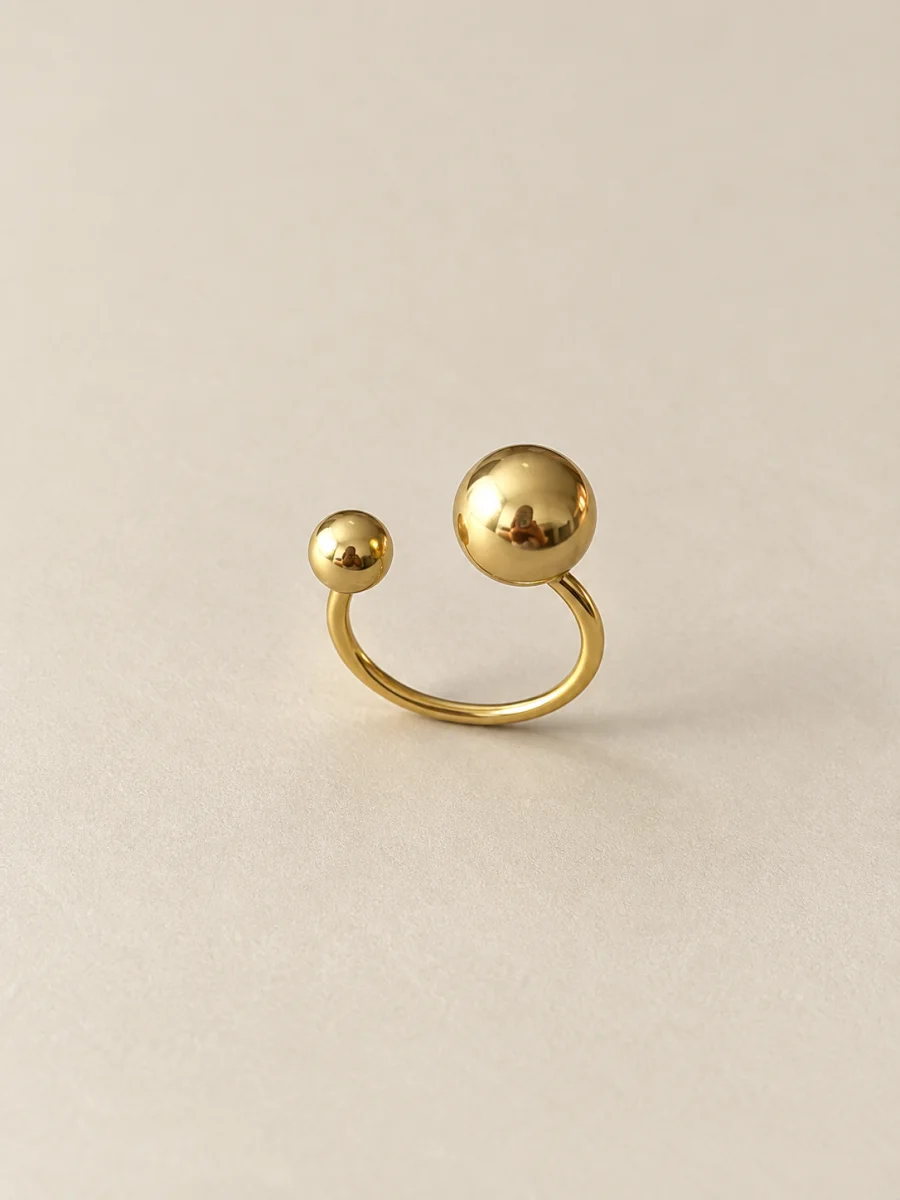 Anel dourado aberto com duas esferas de tamanhos diferentes em fundo bege