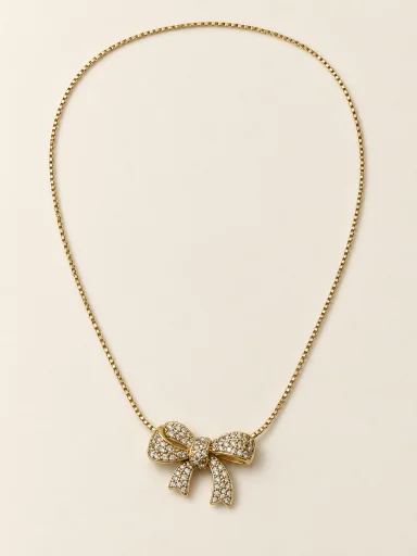 Colar dourado com laço cravejado sobre fundo bege