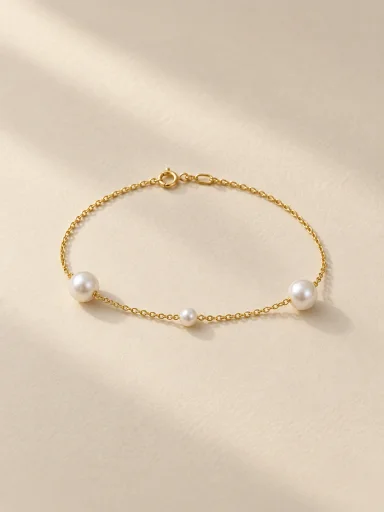 Pulseira dourada de três pérolas sobre fundo bege · formato 3:4