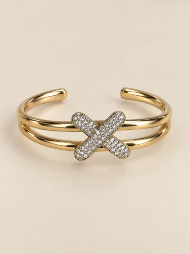 Bracelete dourado com X cravejado sobre fundo bege
